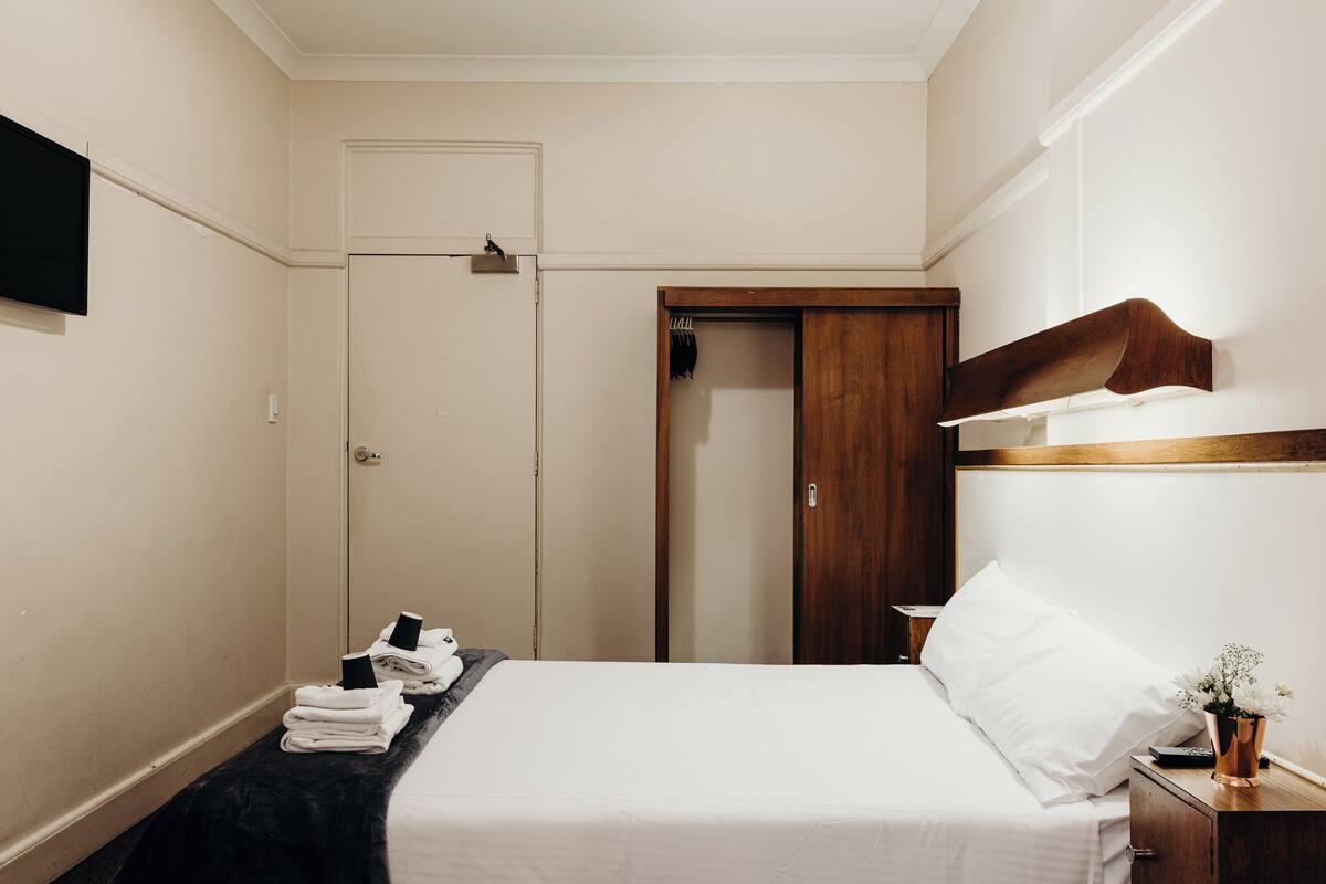Quarto de hotel com cama, mesa de cabeceira com um vaso, guarda-roupa de madeira e porta que dá para outro aposento.