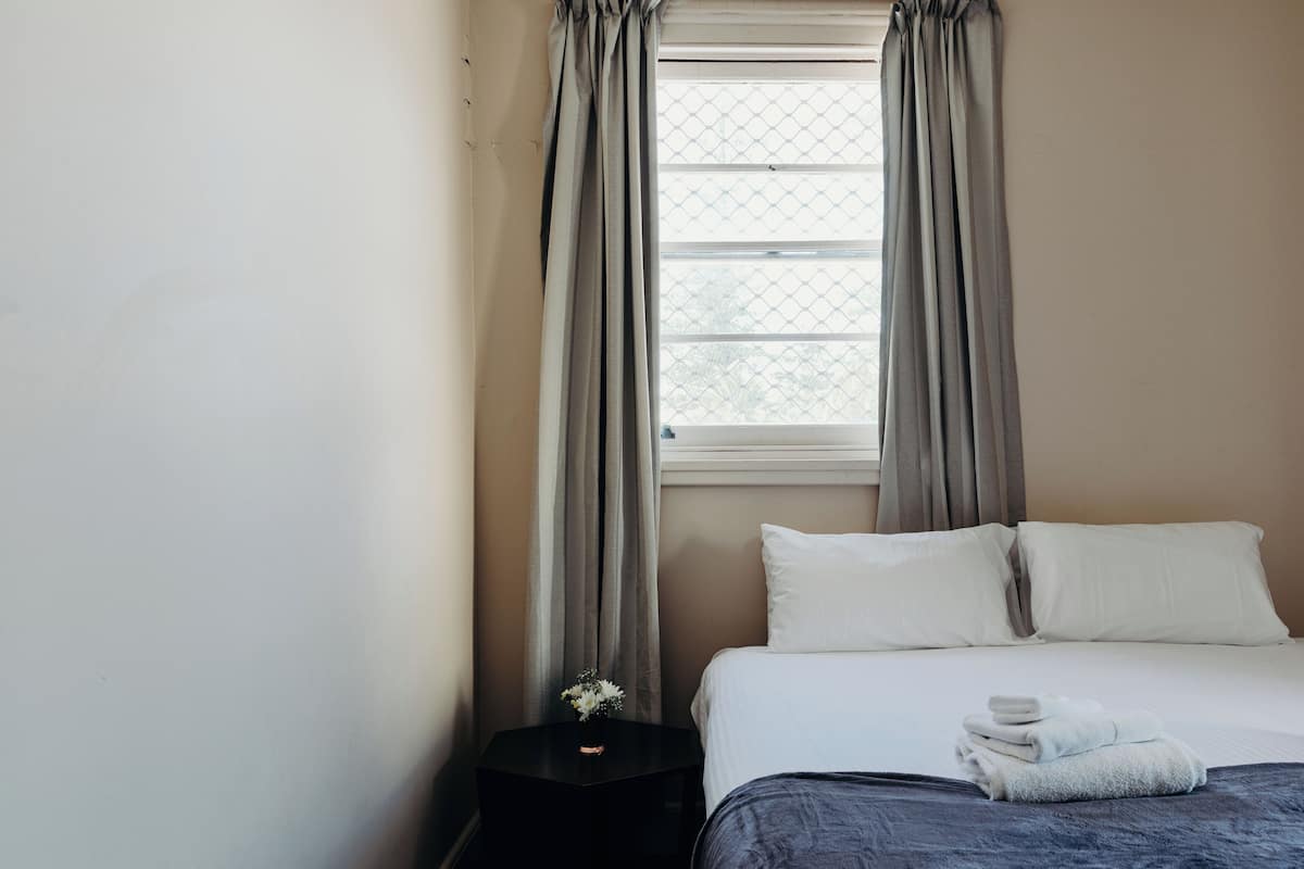 Quarto de hotel com uma cama, almofadas brancas, toalha dobrada e uma pequena mesa com um vaso de flores.