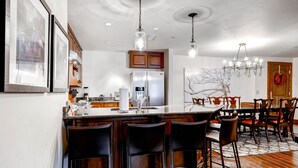 A modern kitchen with a central island, bar stools, and a dining area with wooden chairs and a chandelier. - Ascent by East West Hospitality (Avon)