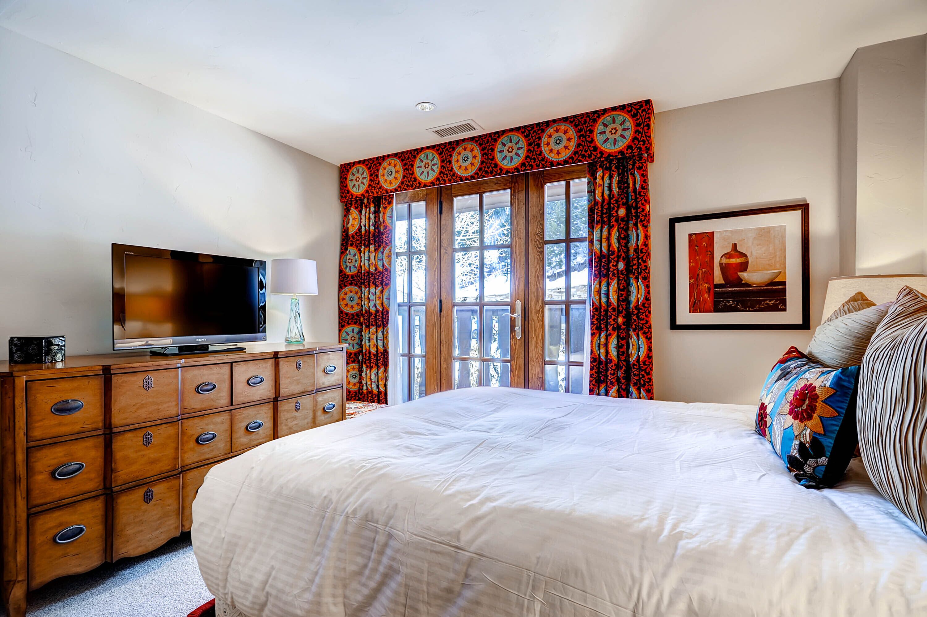 Une chambre à coucher avec un grand lit, une commode en bois, un téléviseur et une fenêtre avec des rideaux.
