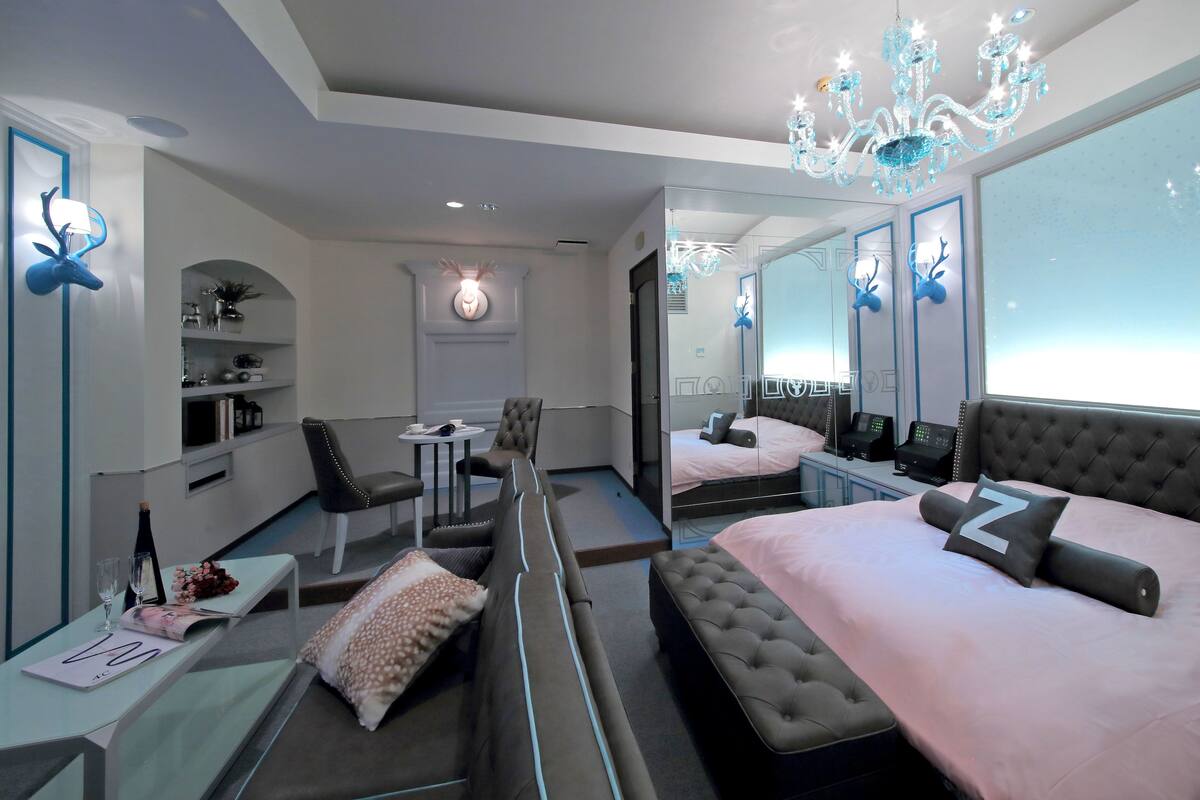 A modern hotel room with a large bed, a seating area with a sofa and chairs, a small table with a vase and glasses, and a chandelier.