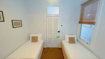 A small, clean room with two single beds, a door, and a window with blinds.