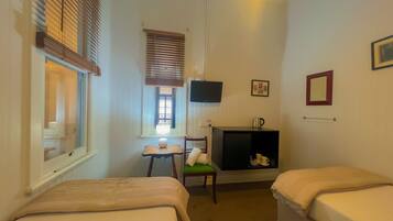 A hotel room with two beds, a small table with a chair, a television, and a shelf with a kettle and cups.
