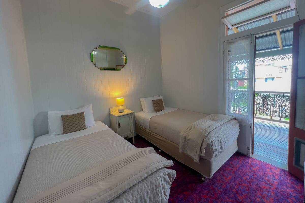 Habitación de hotel con dos camas, una mesita de noche con lámpara, un espejo y una ventana con vistas al exterior.