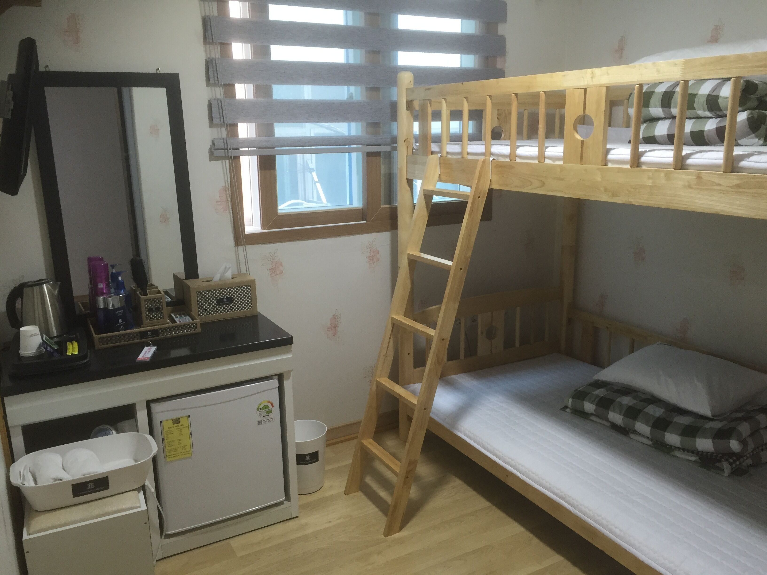 Una cama individual superpuesta con escalera, un pequeño escritorio con artículos de aseo, un refrigerador y una ventana con persianas.