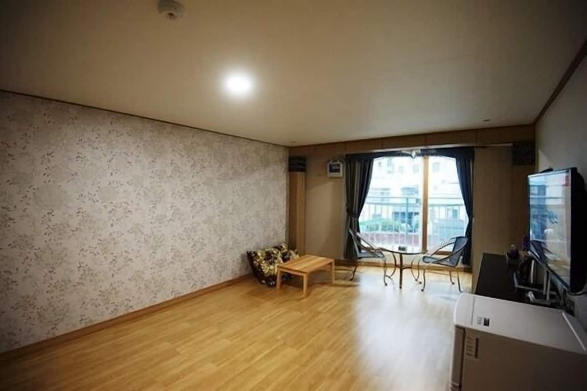 Een ruime woonkamer met houten vloer, een klein tafeltje, een bank en een groot raam met gordijnen.
