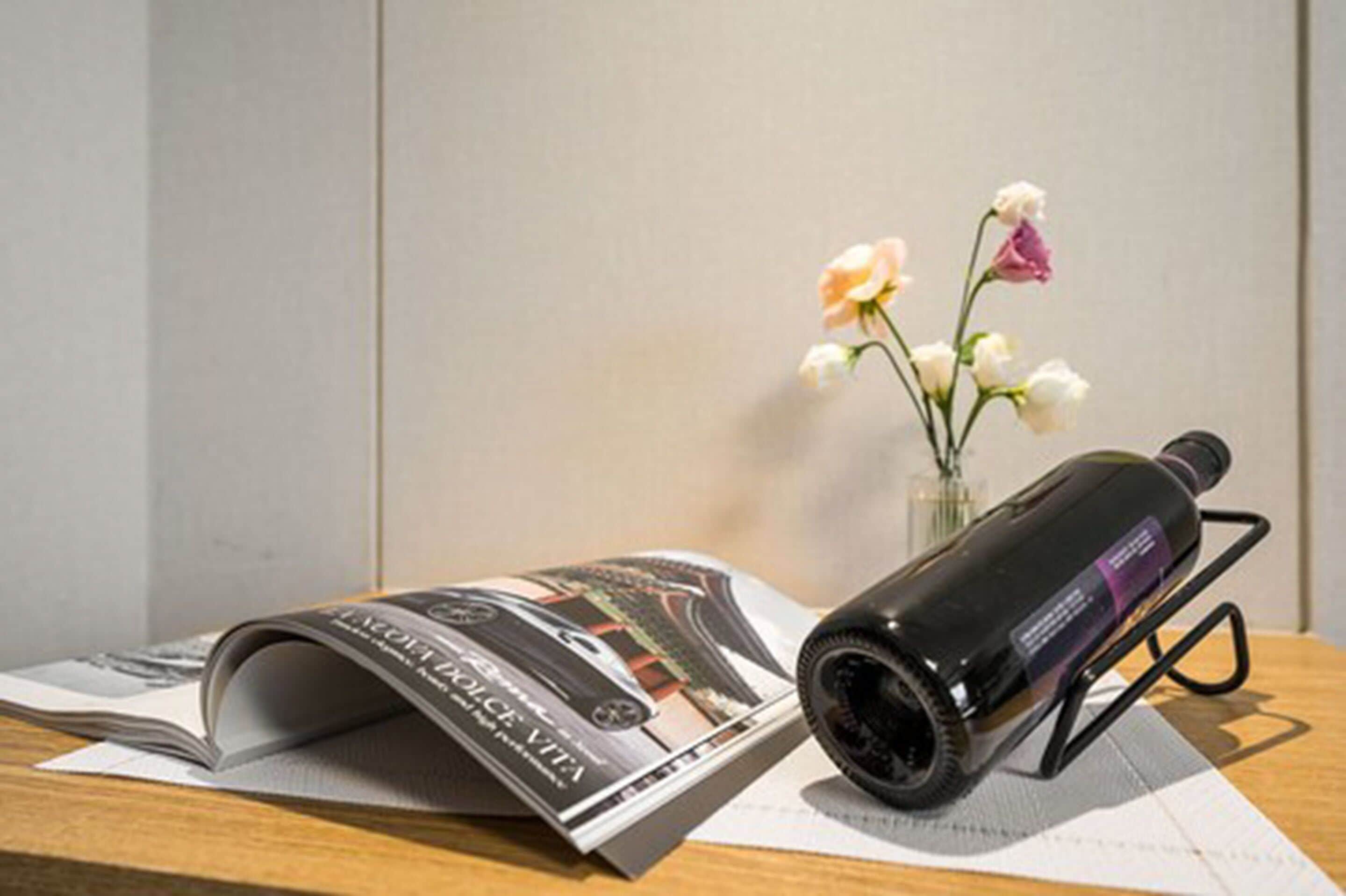A magazine, a bottle of wine, and a vase with flowers on a wooden table.