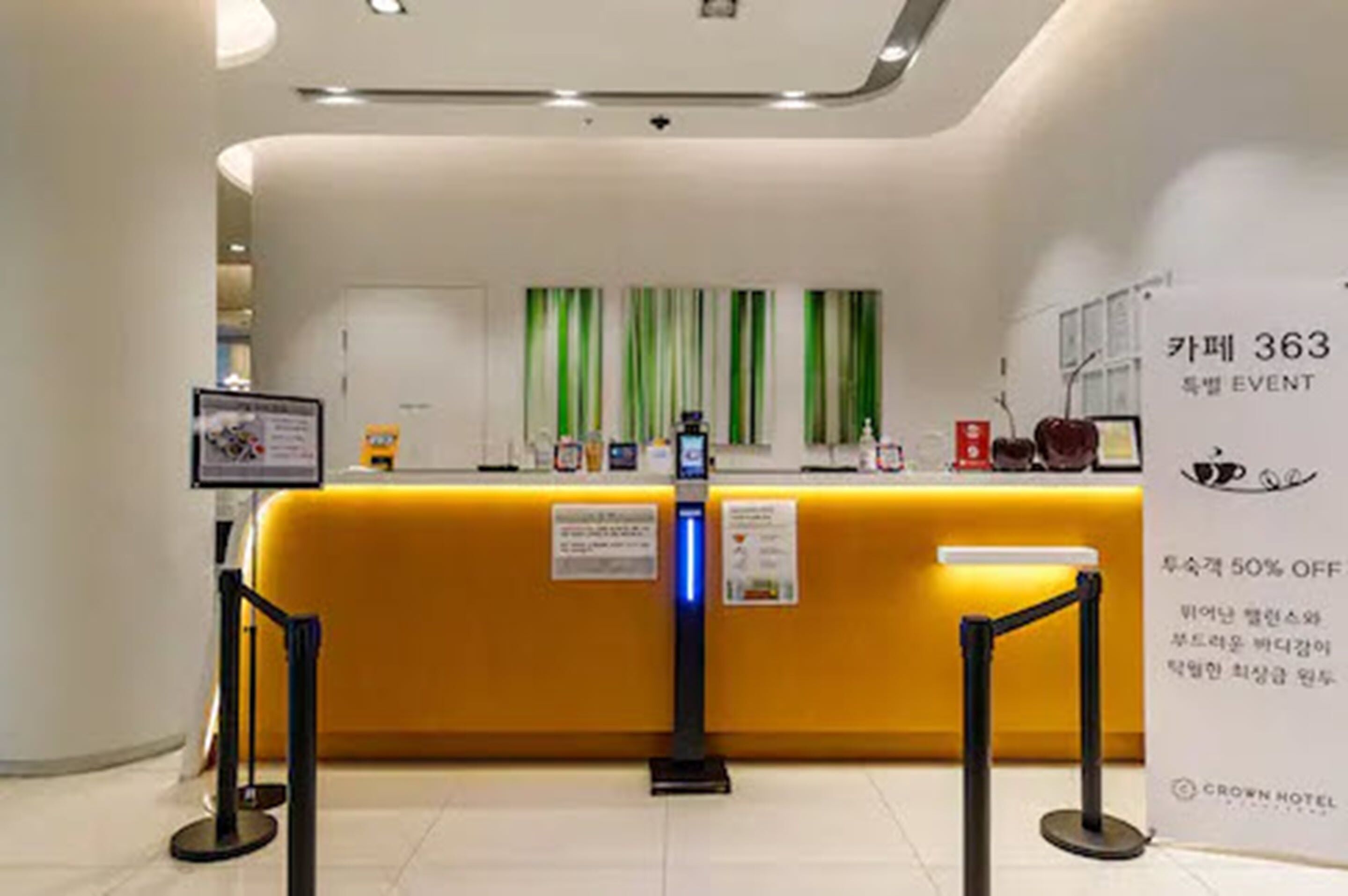 Ein Hotel-Rezeptionsbereich mit einem gelben Empfangstresen, einer digitalen Check-in-Kiosk und einem Schild, das auf ein 50%-Rabatt-Event hinweist.