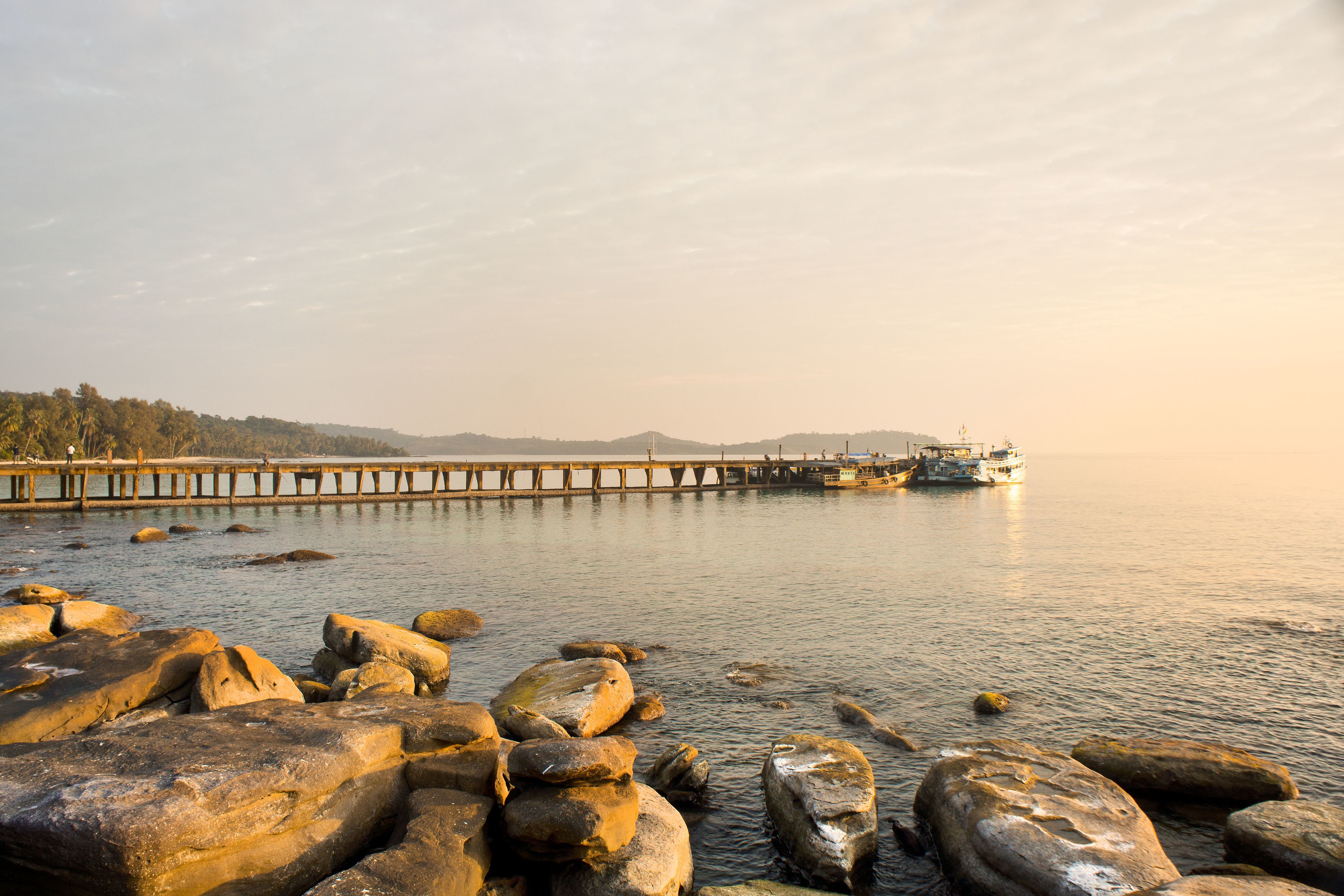A rocky shoreline with a pier extending into calm waters, a boat docked at the end, and distant hills.