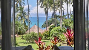 Habitación con vistas a un paisaje tropical, que incluye palmeras y una playa. En primer plano, hay dos sillas blancas con cojines y una mesita.