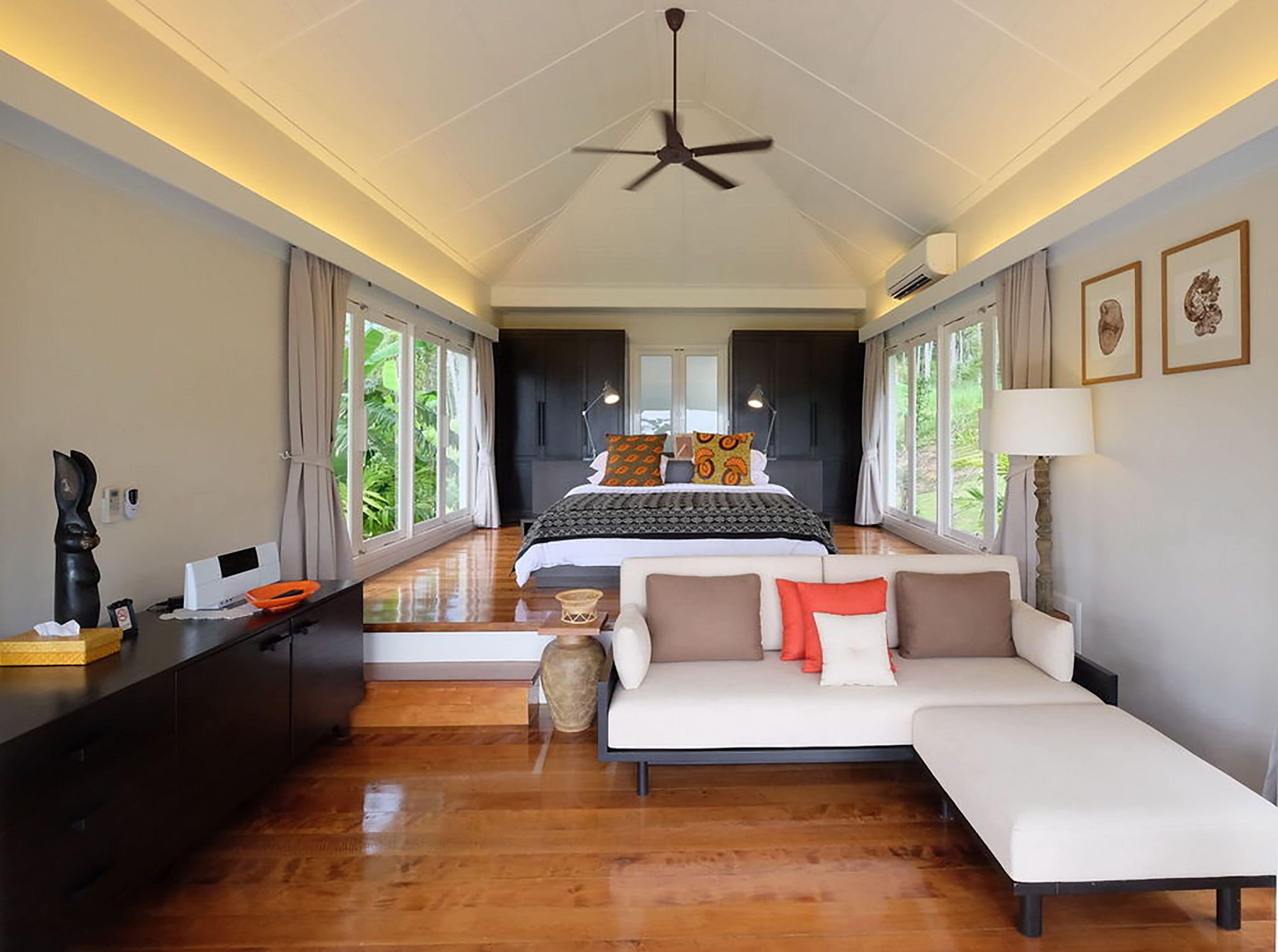 A spacious bedroom with a large bed, a sofa, and a desk. There are large windows providing natural light and a view of greenery outside.