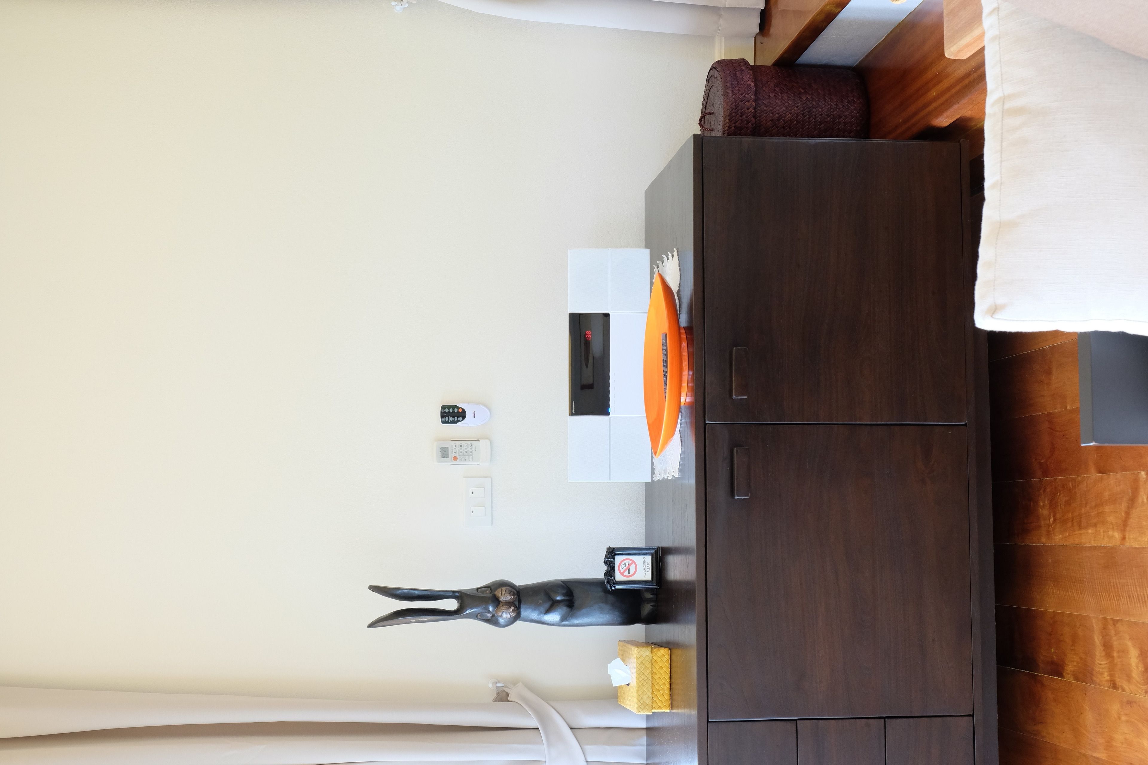 A bedroom with a wooden wardrobe, a bed with a white pillow, a bedside table with a remote control, a tissue box, and a decorative item.
