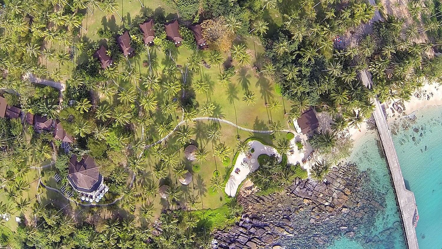 Vue aérienne d’un complexe hôtelier tropical, avec des palmiers, une plage et une côte rocheuse.