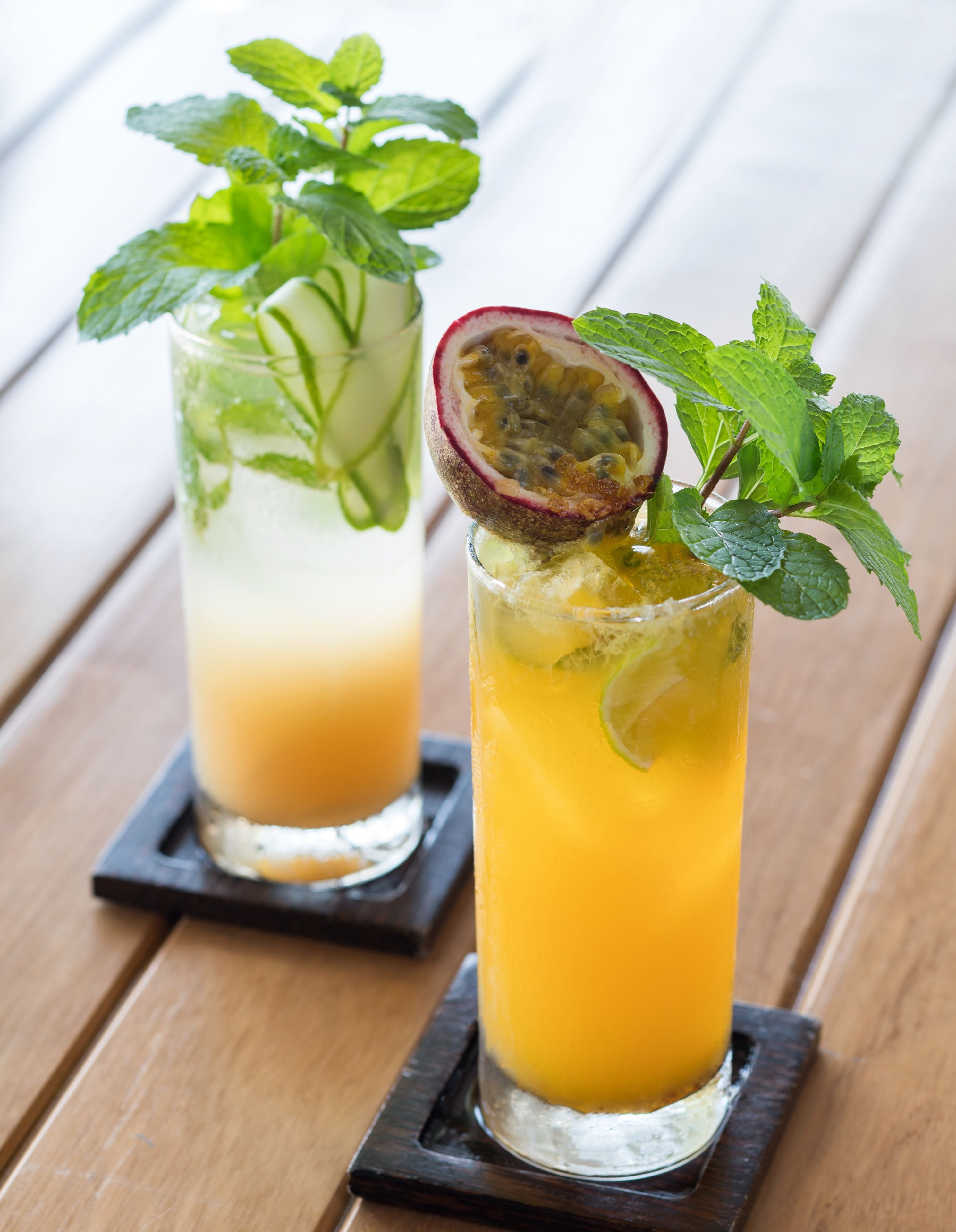 Two tall glasses of a yellow beverage with mint leaves and a passion fruit slice, placed on wooden coasters.