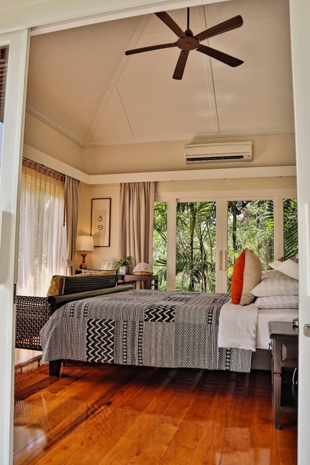 Ein Schlafzimmer mit einem Bett, einem Deckenventilator, einer Klimaanlage, einem Fenster mit Vorhängen und Blick ins Grüne.