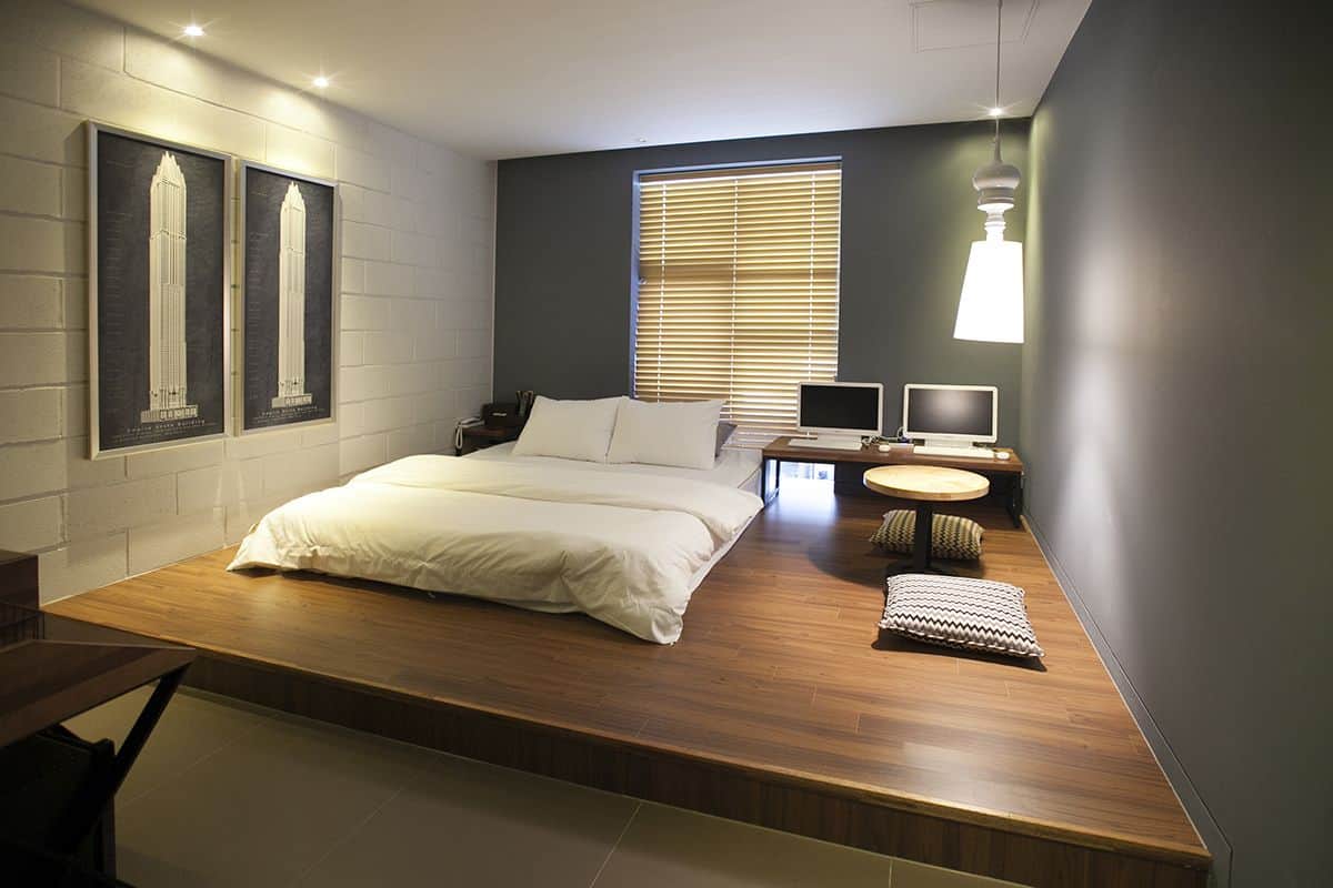 Un dormitorio moderno con cama, escritorio, dos monitores y ventana con persianas.