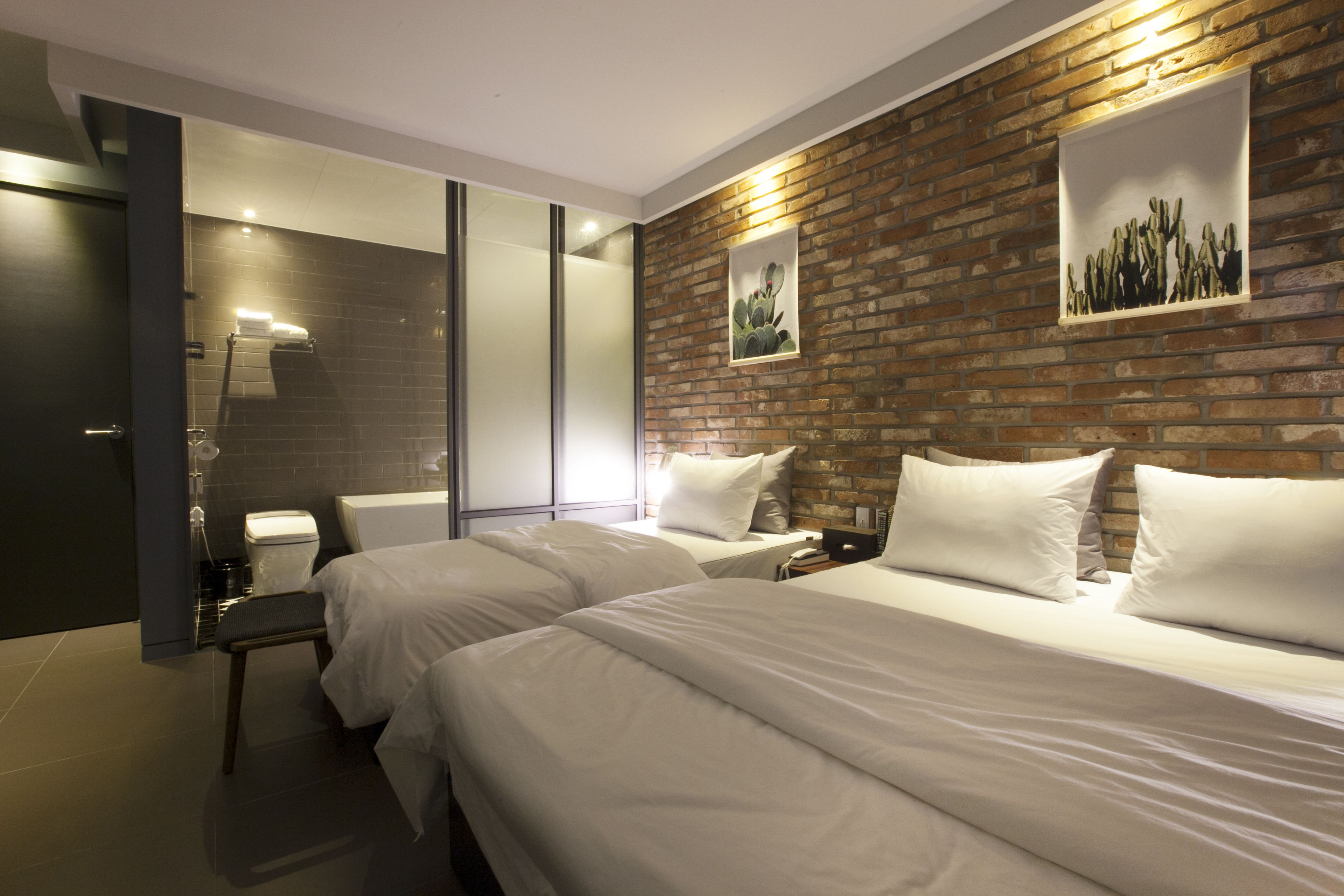 A hotel room with two beds, a brick accent wall, a framed cactus artwork, and a modern bathroom with a glass shower enclosure.