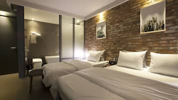A hotel room with two beds, a brick accent wall, a framed cactus artwork, and a modern bathroom with a glass shower enclosure.
