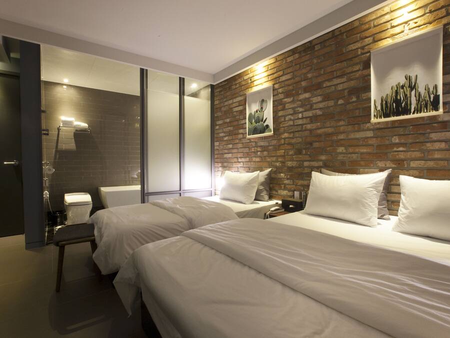 A hotel room with two beds, a brick accent wall, a framed cactus artwork, and a modern bathroom with a glass shower enclosure.