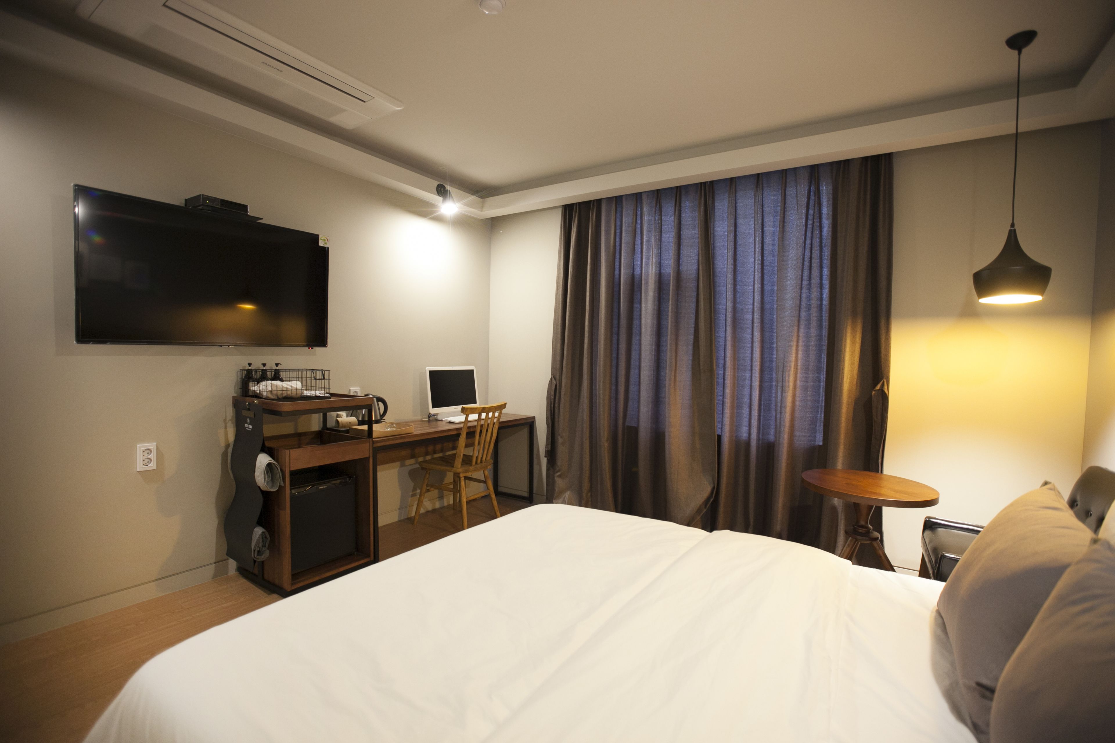 Quarto de hotel com cama, escrivaninha, cadeira, televisão e cortinas.