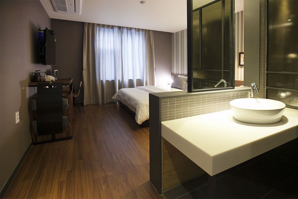 Ein modernes Hotelzimmer mit einem großen Bett, einem integrierten Schreibtisch, einem großen Fenster mit Vorhängen und einem modernen Badezimmer mit großem Spiegel und Waschbecken.