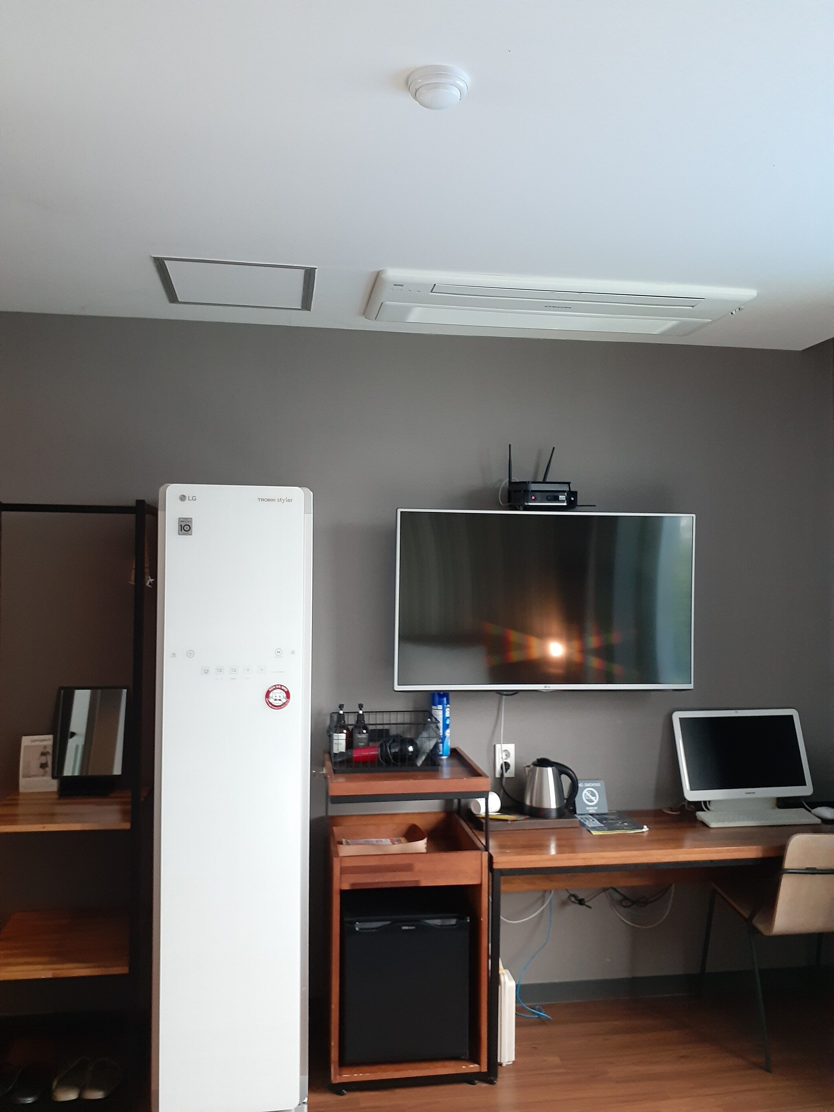 A hotel room with a flat-screen TV mounted on the wall, a small refrigerator, a desk with a laptop, and a compact refrigerator.