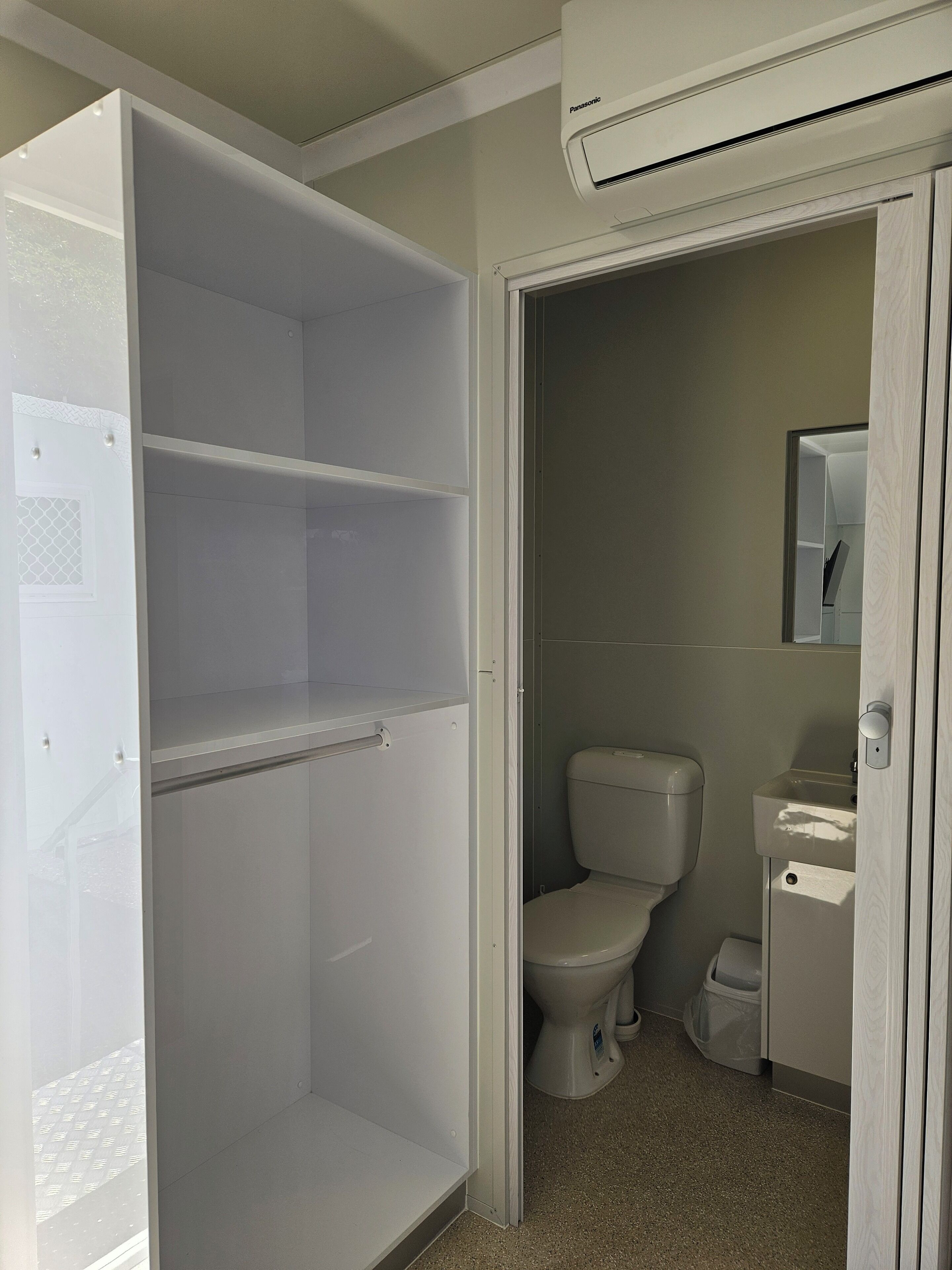 A bathroom with a toilet, a sink, and a mirror. There is a white cabinet with a door, and an air conditioning unit above.