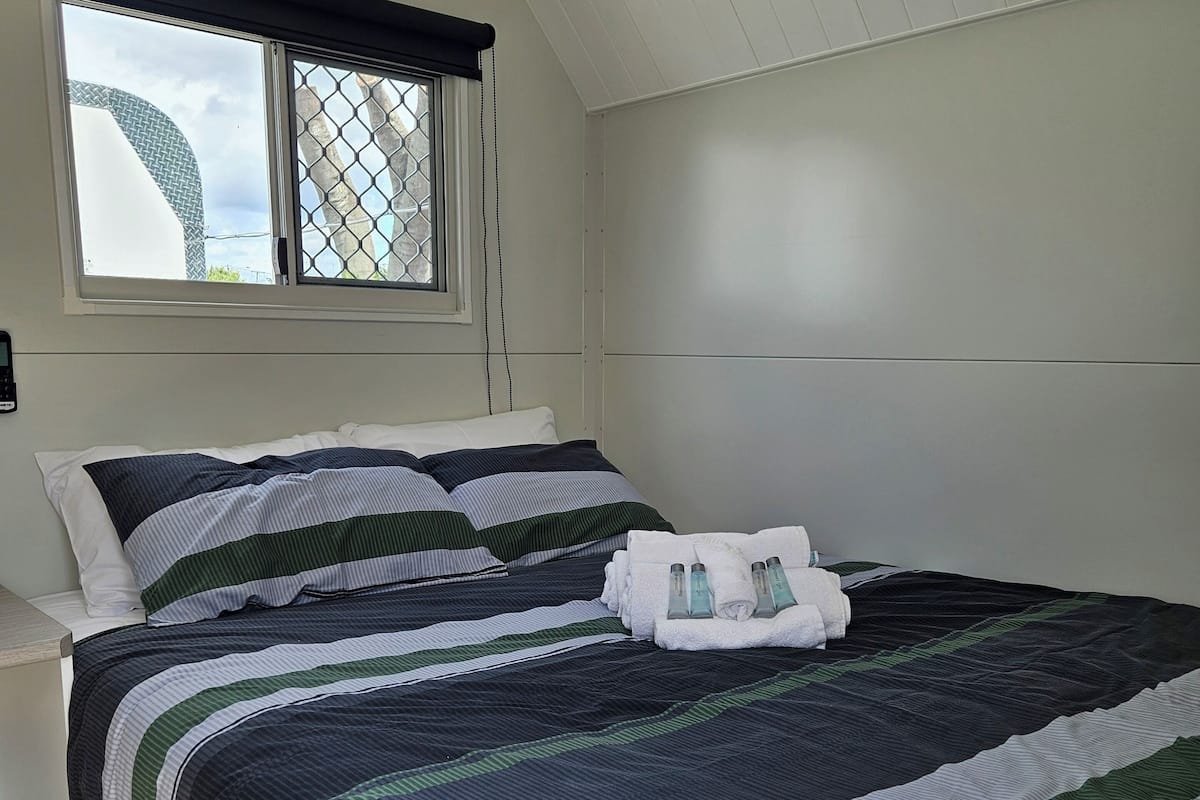 Una cama bien tendida con ropa de cama a rayas, una ventana con mosquitero y un cabecero blanco.
