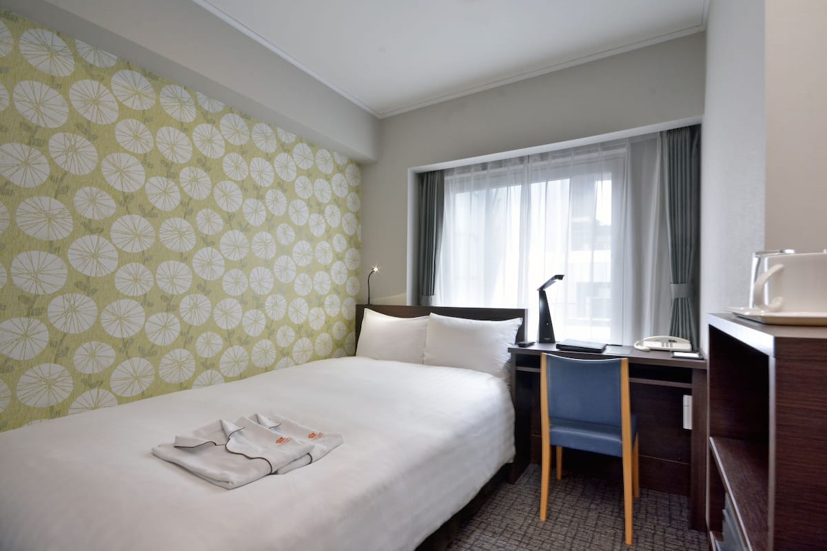 A hotel room with a neatly made bed, a desk with a chair, a lamp, and a window with curtains.