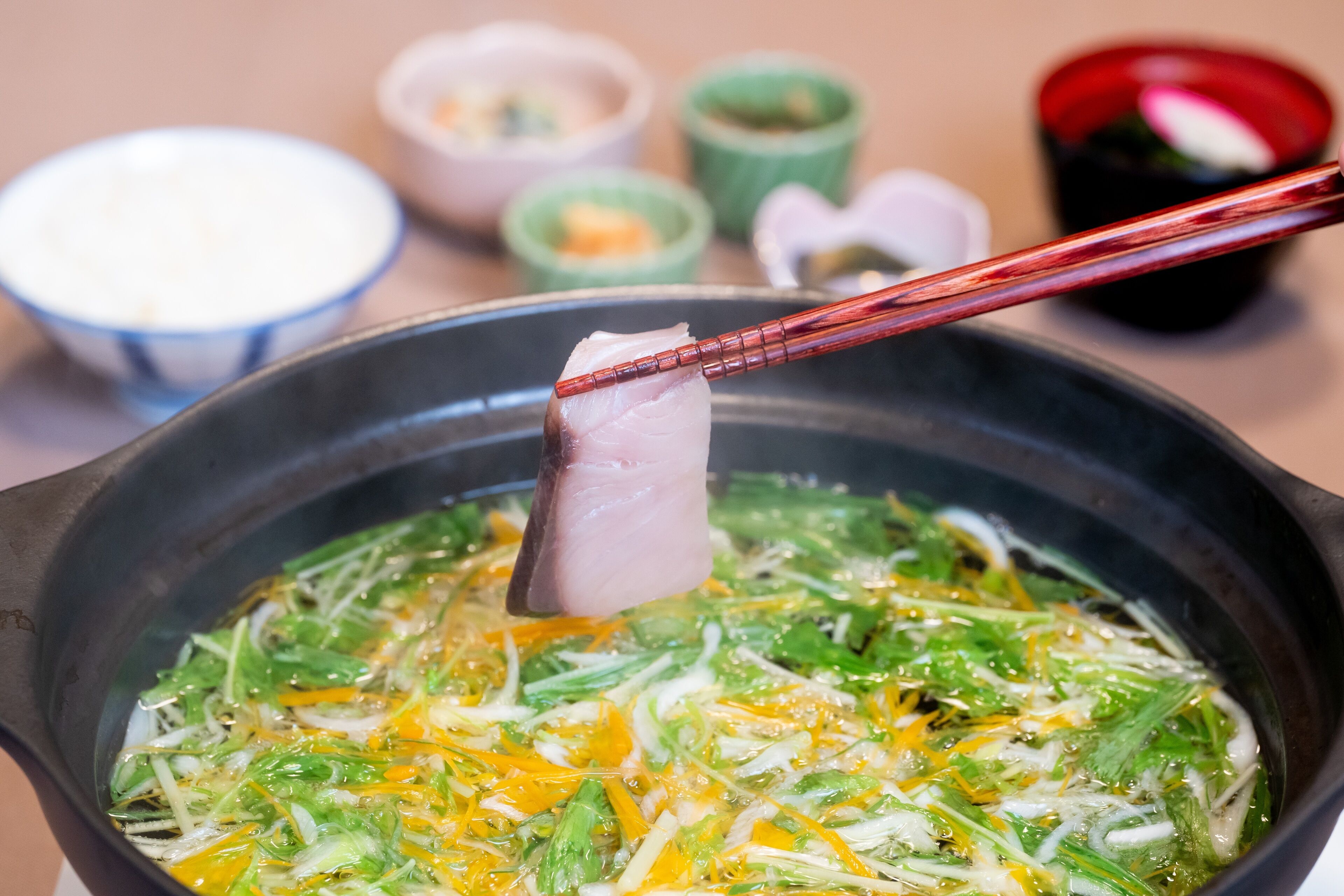 A close-up of a dish with various vegetables and egg in a pot, with chopsticks lifting a piece of meat.
