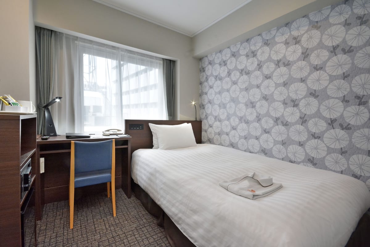 A hotel room with a bed, a desk, a chair, a lamp, and a window with curtains.