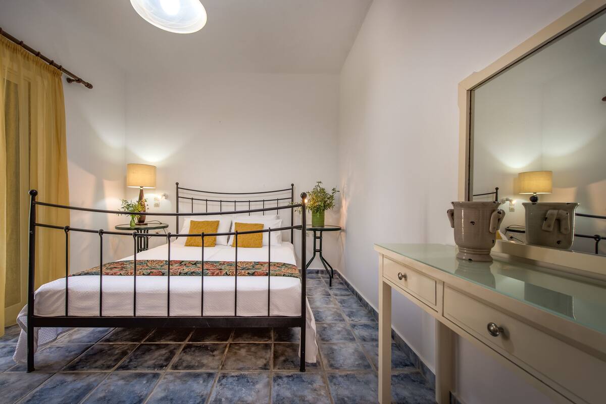 A bedroom with a metal bed frame, a bedside table with a lamp, a dresser with a mirror, and a potted plant.