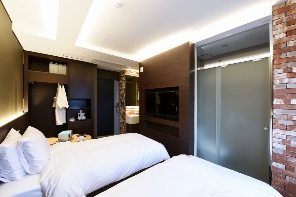 A hotel room with two beds, a wardrobe, a TV, and a small table with a cup and a book.