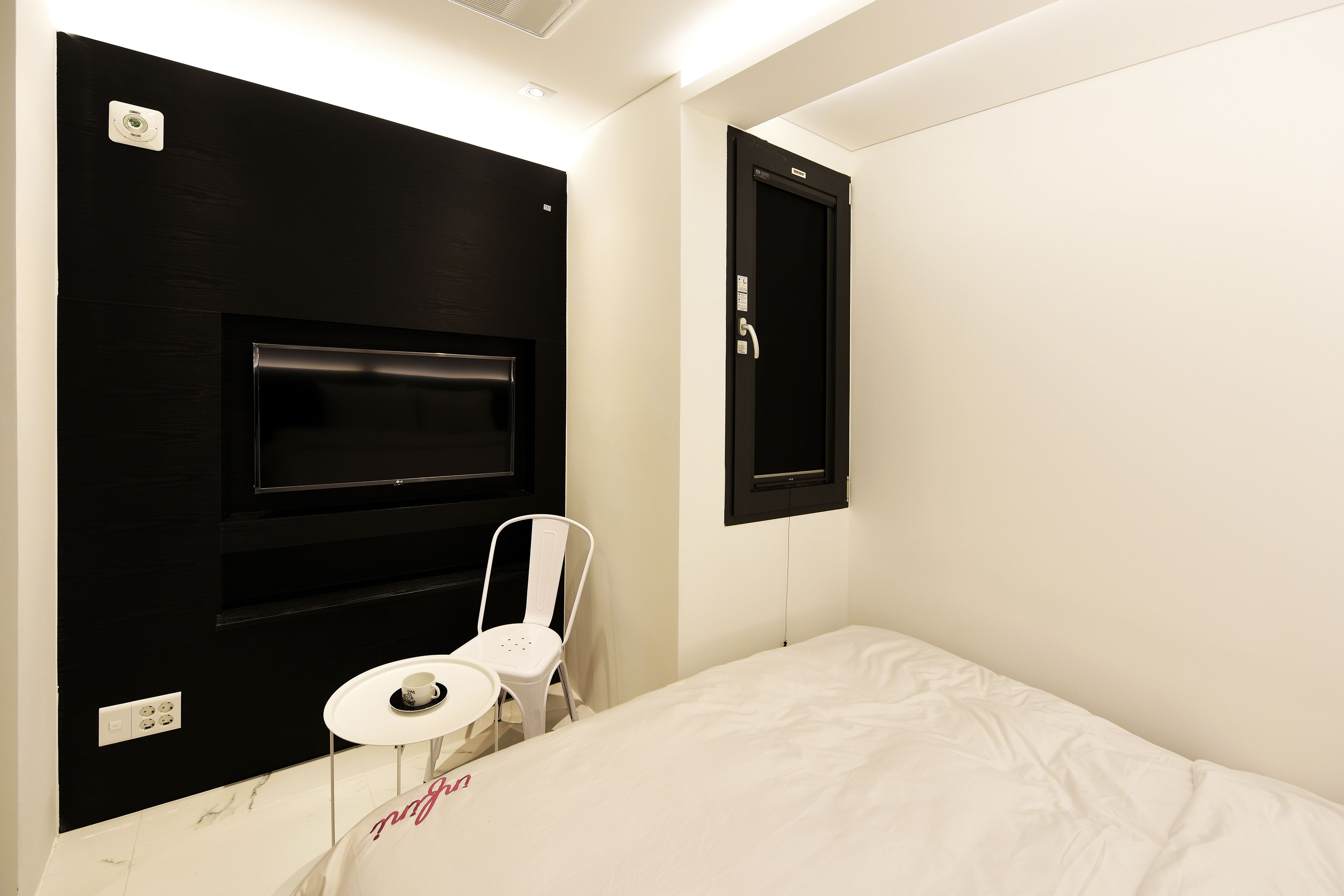 A hotel room with a bed, a small white table, a white chair, a black wall-mounted TV, and a black door.