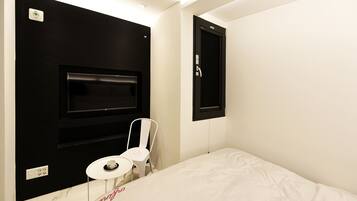 A hotel room with a bed, a small white table, a white chair, a black wall-mounted TV, and a black door.