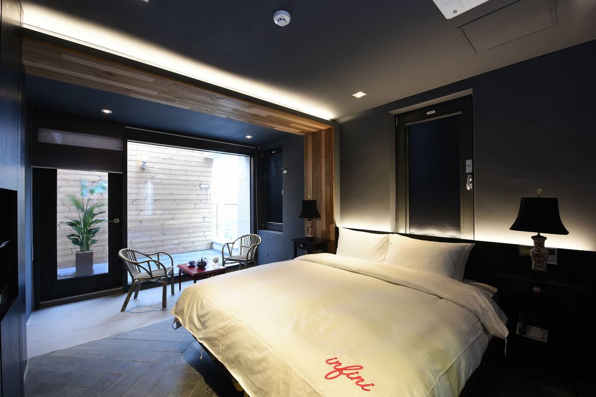 A modern hotel room with a large bed, a small table, two chairs, a window with a view, and a nightstand with a lamp.