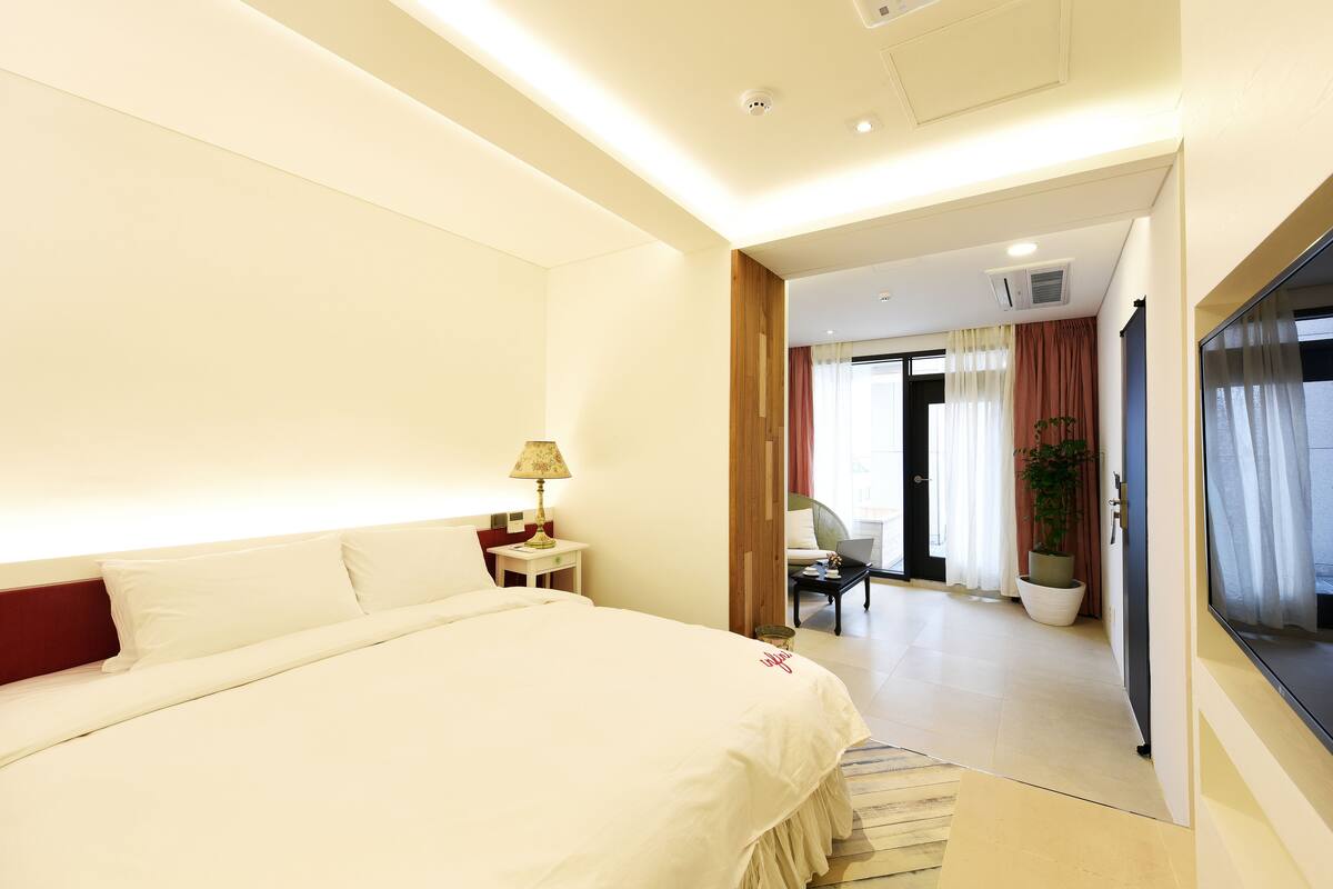 A hotel room with a large bed, a bedside table with a lamp, a chair, a potted plant, and a view of the outside through a door.