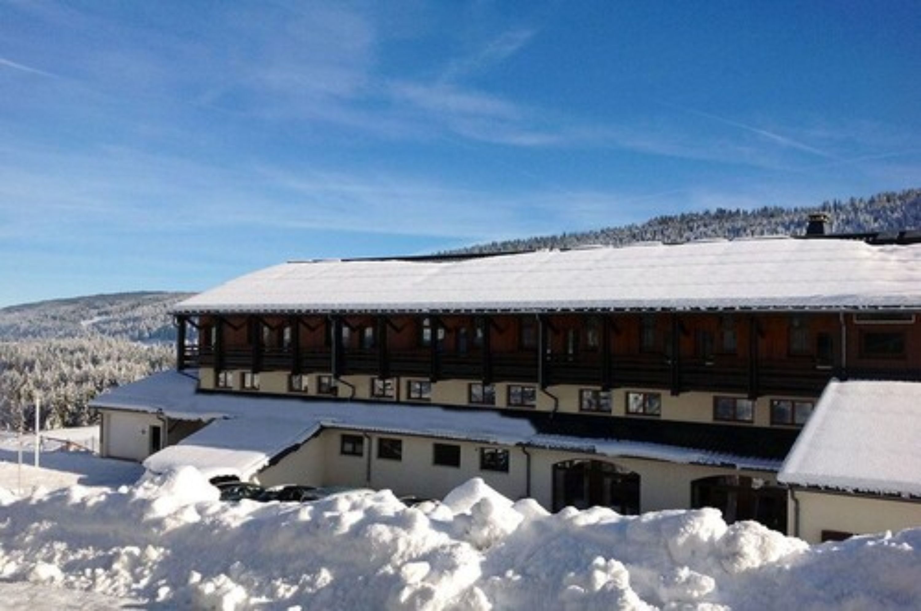 Un edificio tradizionale in legno con tetto imbiancato dalla neve, immerso in un paesaggio innevato e con una collina boscosa sullo sfondo.