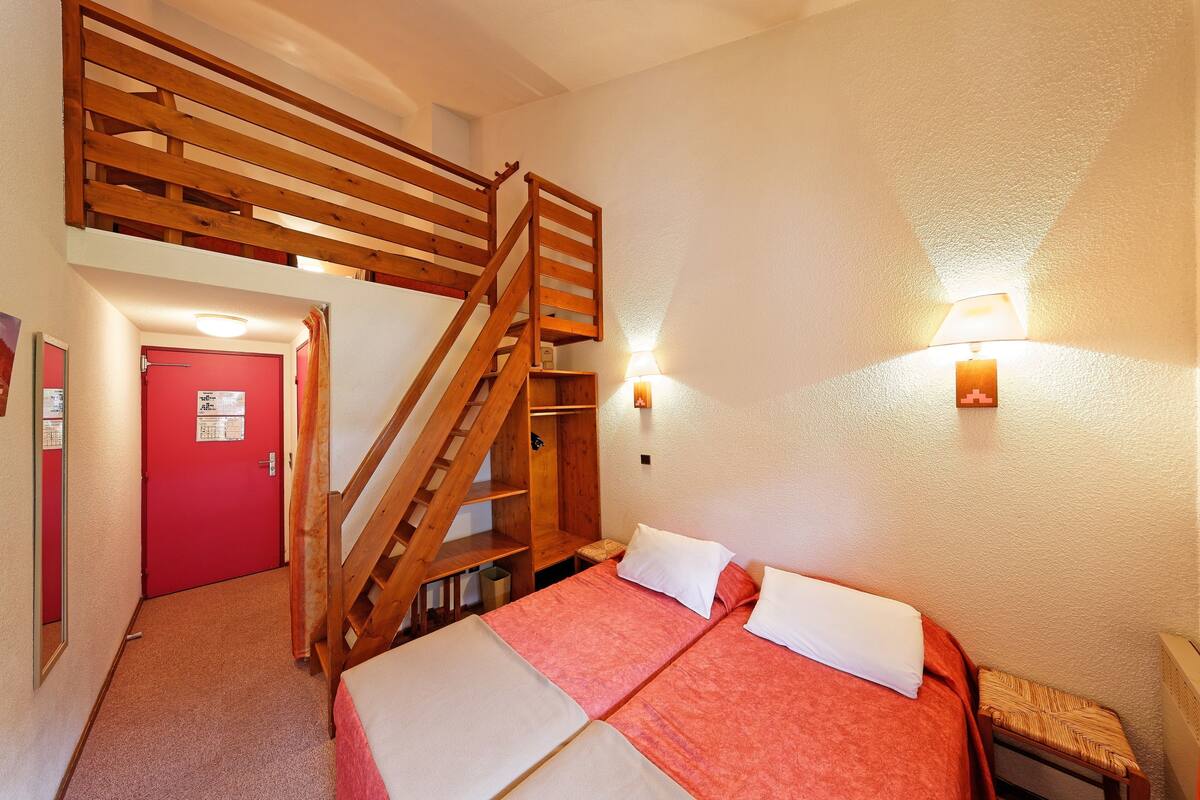 A room with two beds, a wooden staircase, a red door, and a wall-mounted lamp.