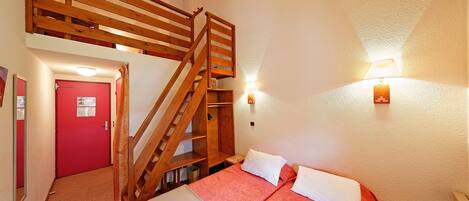 A room with two beds, a wooden staircase, a red door, and a wall-mounted lamp.