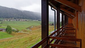 A wooden balcony with a view of a rural landscape, including houses and a forested hill.