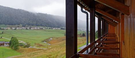 Un balcón de madera con vista a un paisaje rural, que incluye casas y una colina boscosa.