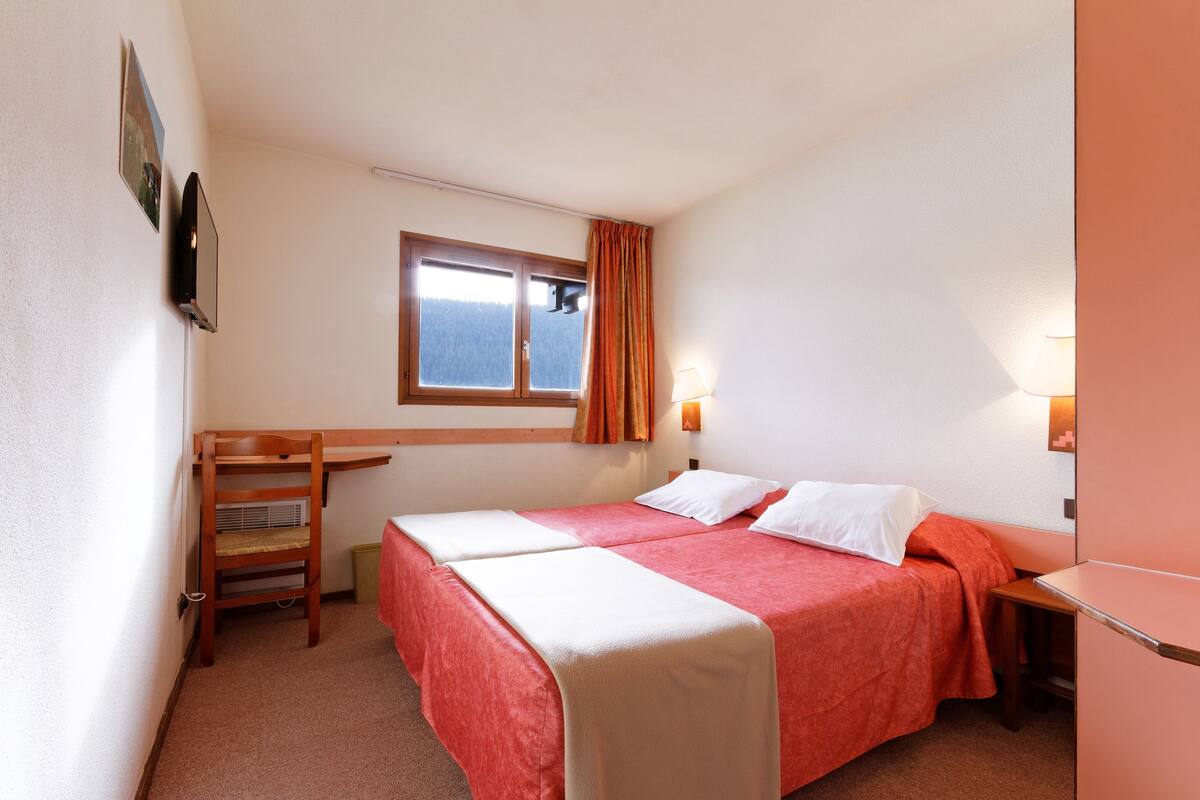 A small, well-lit room with two single beds, a desk, a chair, and a window with curtains.