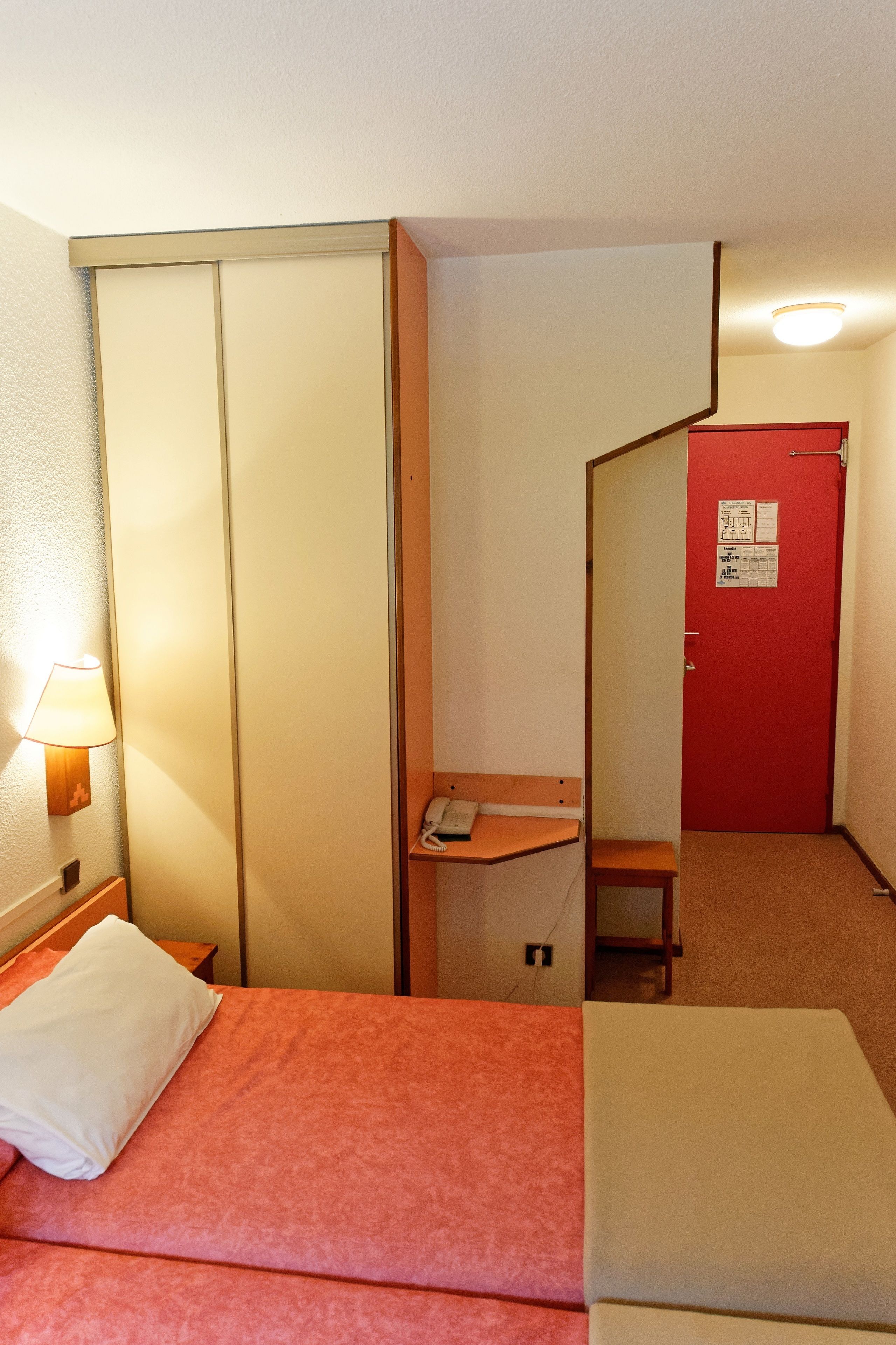 Quarto de hotel com uma cama, uma mesa de cabeceira com abajur, uma porta vermelha e um pequeno banco de madeira.