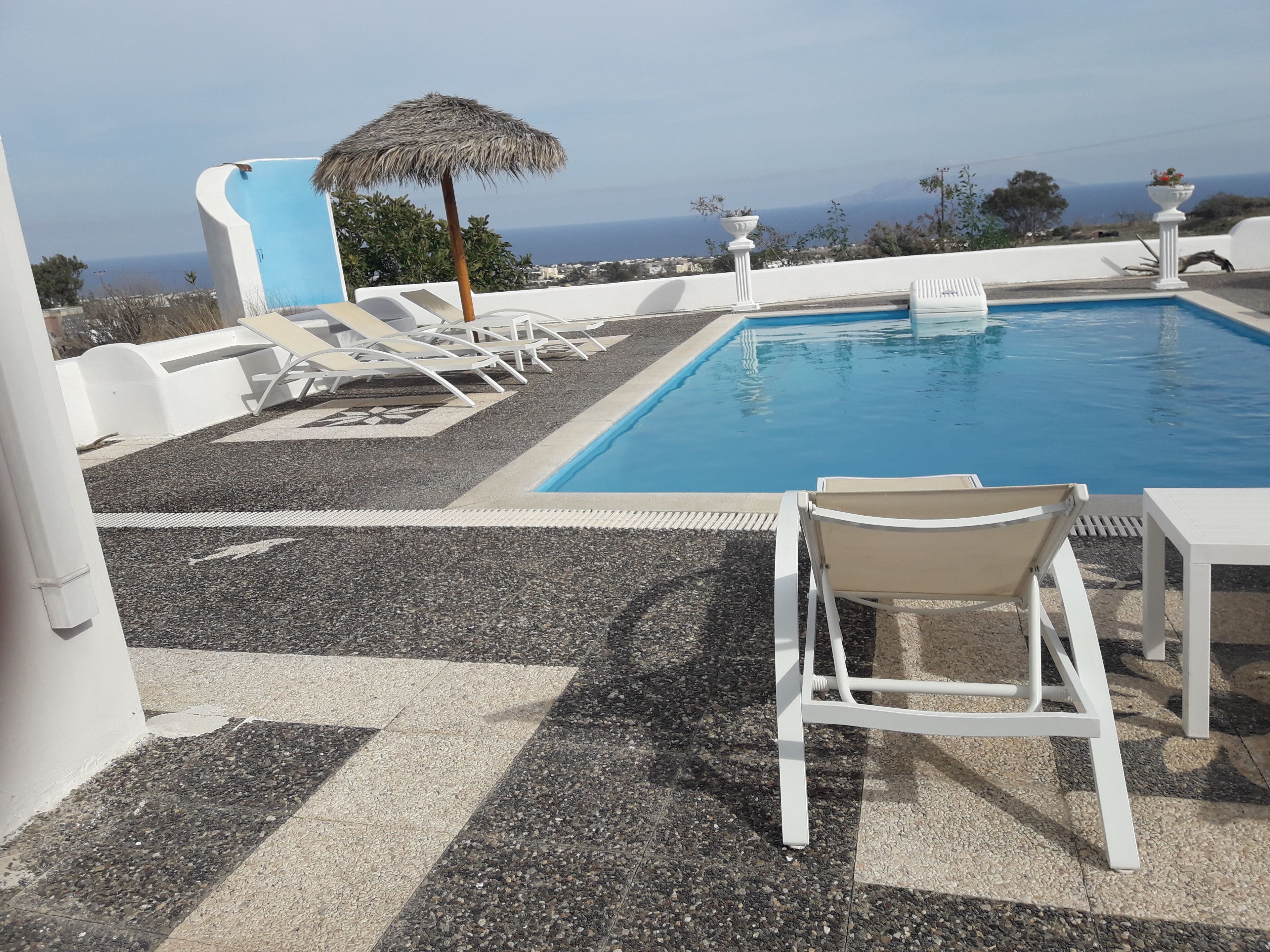Un área de piscina con sillas de descanso y una mesa, rodeada por un piso de adoquines y paredes blancas.