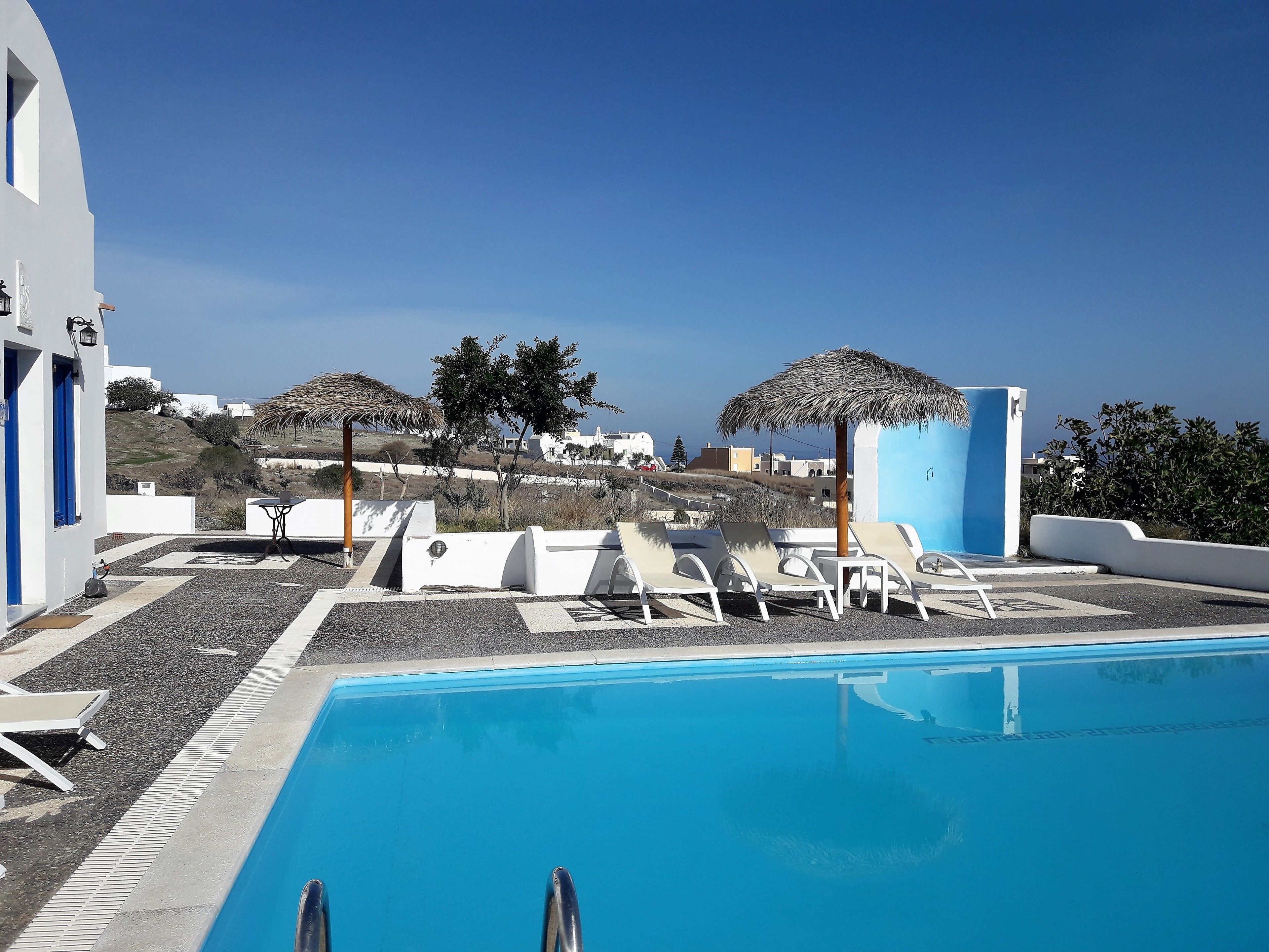 A pool area with lounge chairs and umbrellas, a clear blue sky, and white buildings in the background.