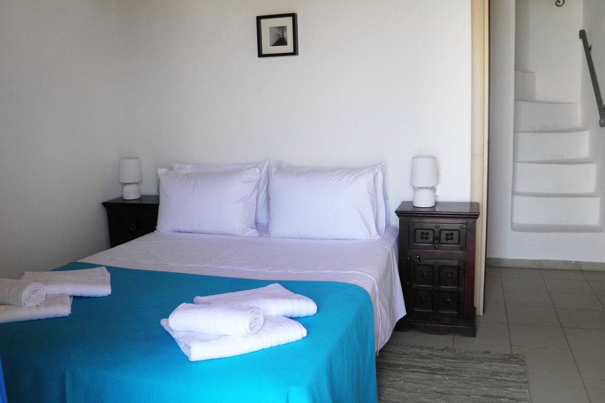 A bed with white linens, a blue bedspread, and two white towels on the bed.