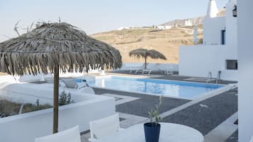 Un área junto a la piscina con una sombrilla de paja, sillas reclinables blancas y una pequeña mesa con una planta en maceta.