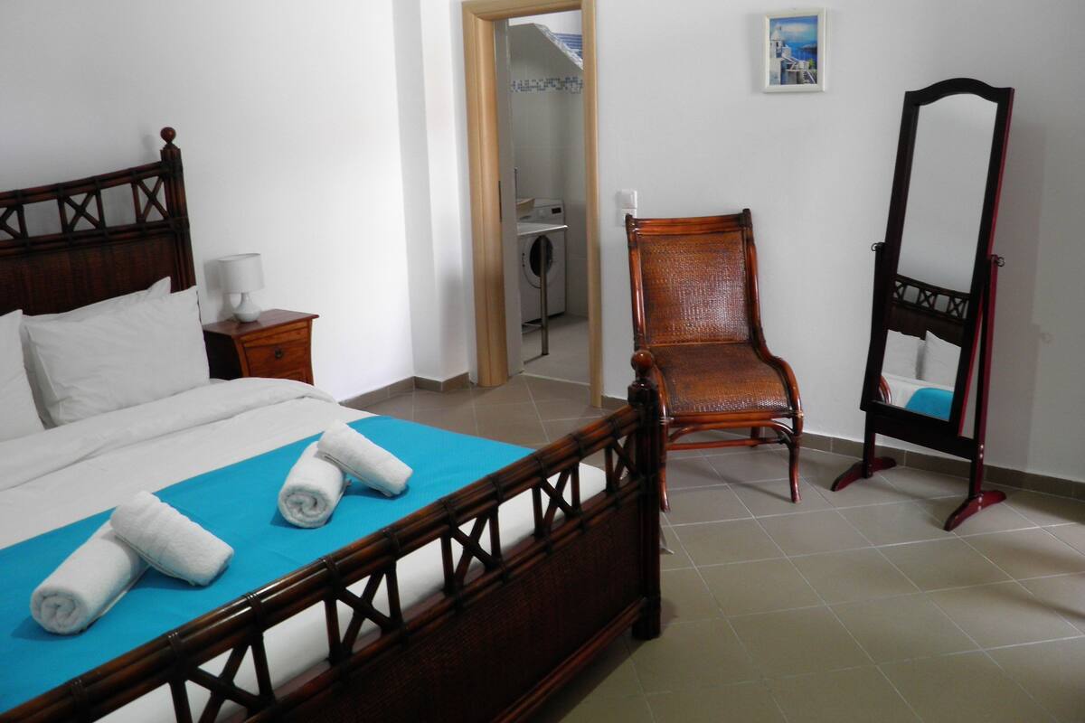 A bedroom with a wooden bed, a chair, a mirror, and a small table with a lamp.