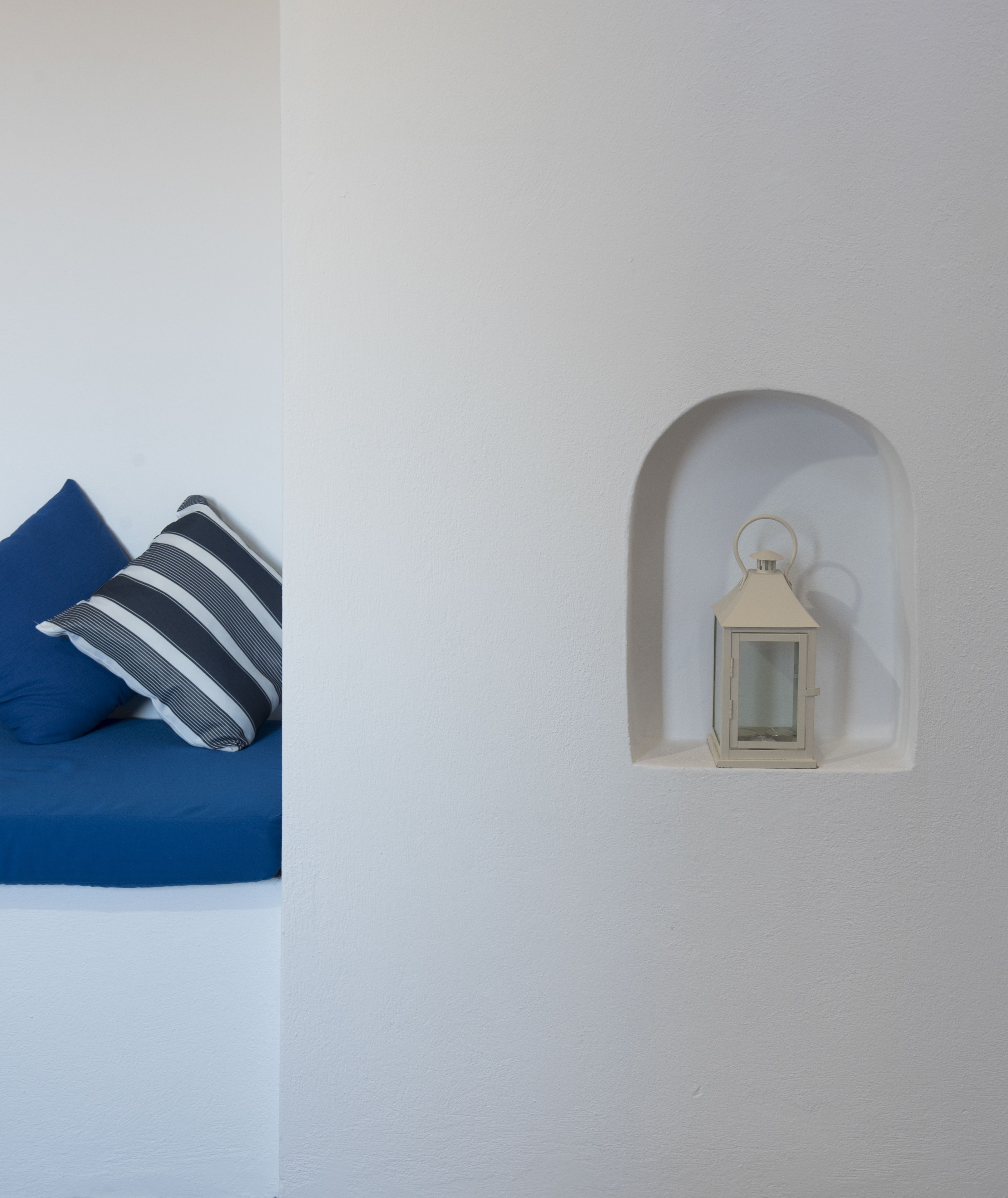A white wall with a small, arched window niche containing a lantern. A blue sofa with a striped pillow is visible to the left.